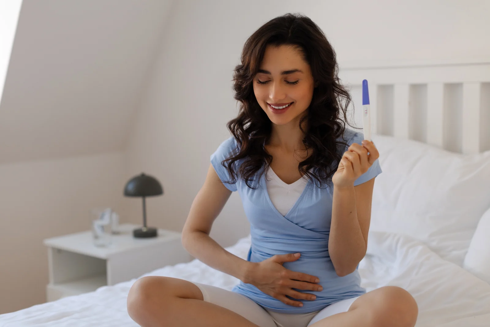 Pregnant woman smiling while holding a positive pregnancy test on a bedroom bed